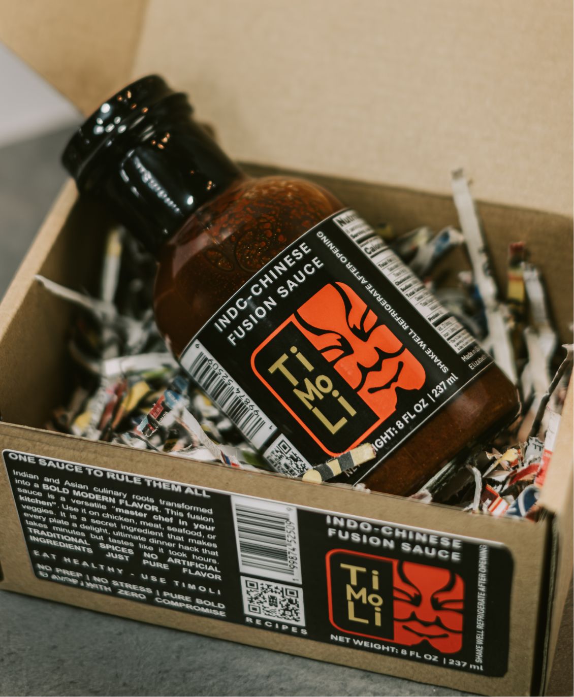 A bottle of Ti Mo Li Indo-Chinese Fusion Sauce rests in a box filled with shredded paper, showcasing its bold black and red label featuring a stylized face logo. The box has label text and branding details.