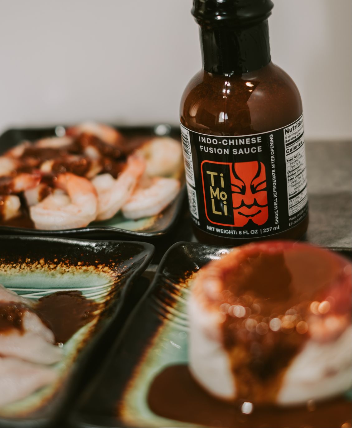 A bottle of TiMiLi Indo-Chinese fusion sauce is surrounded by plates of shrimp and other food, each topped with the dark sauce. The bottle label features bold graphics and text.