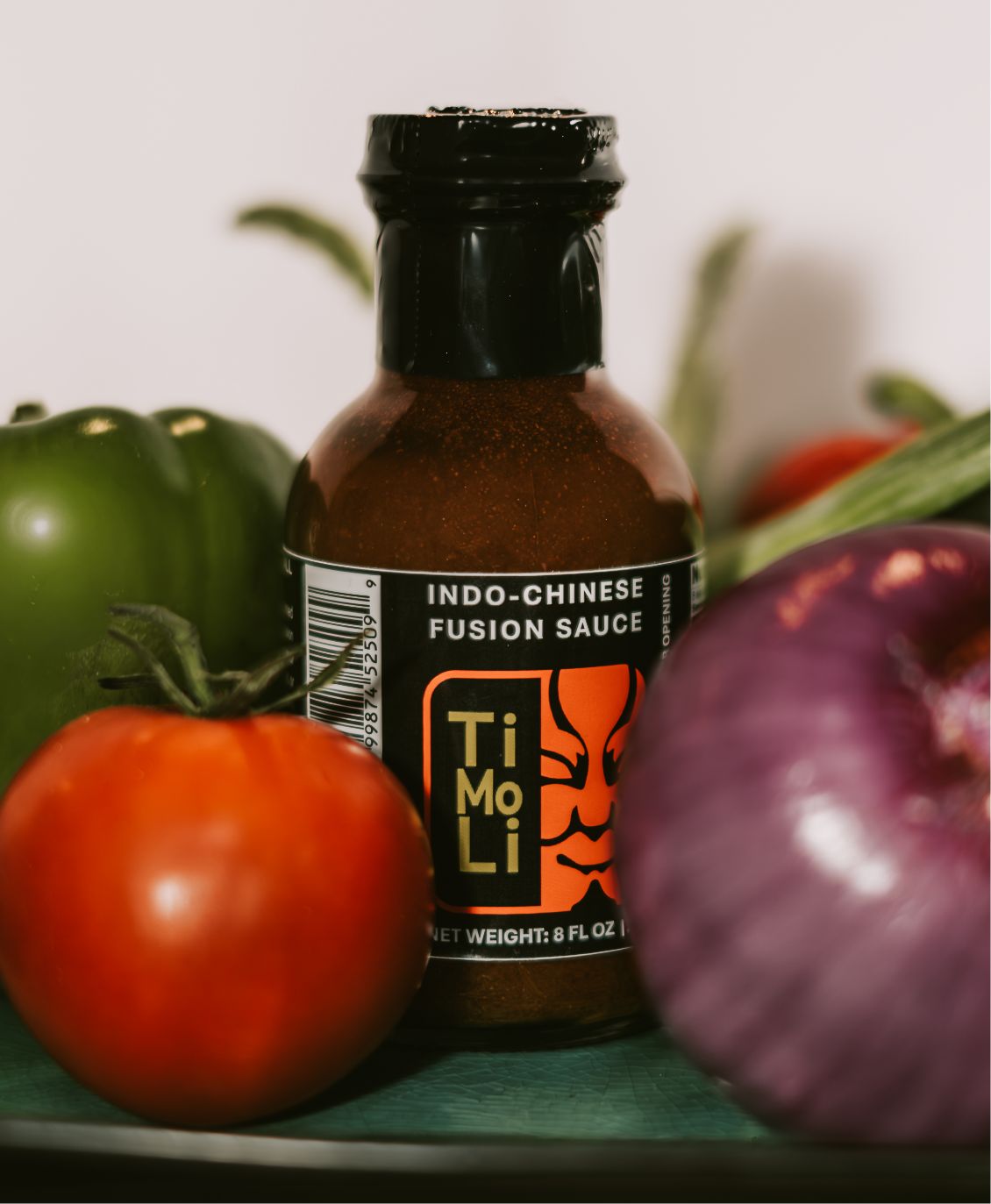 A bottle of TiMoLi Indo-Chinese fusion sauce is surrounded by a green bell pepper, a tomato, an onion, and other fresh vegetables on a green surface.