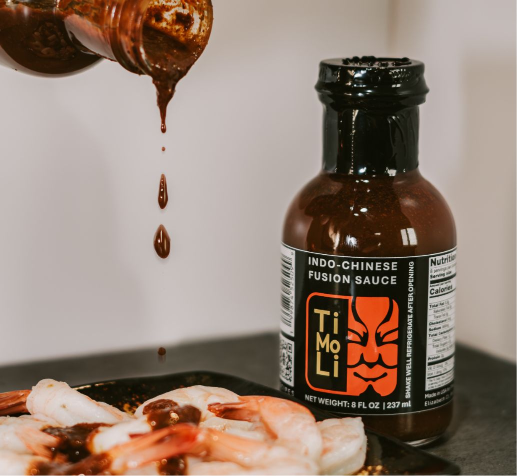 A bottle of TiMoLi Indo-Chinese Fusion Sauce stands on a counter while sauce is being poured over cooked shrimp on a plate nearby.