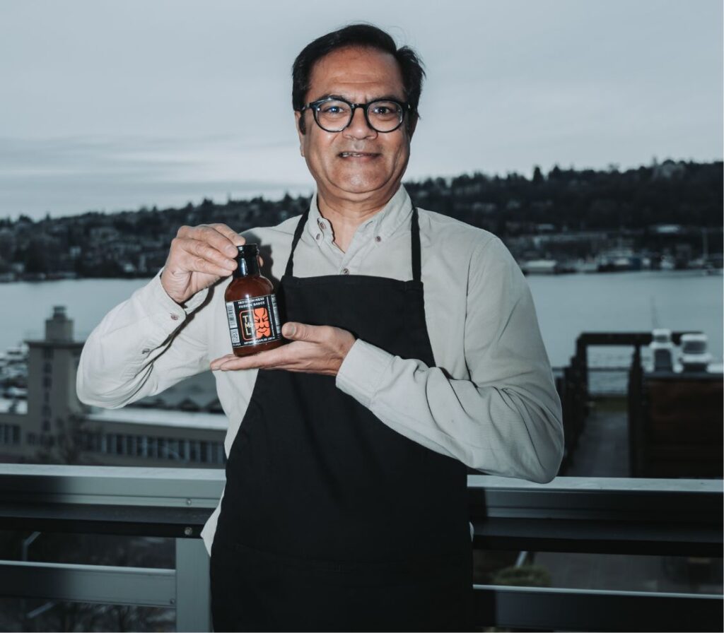 A man wearing glasses and an apron stands on a balcony, smiling and holding a bottle of sauce. The background shows a waterfront scene with buildings, water, trees, and cloudy skies.