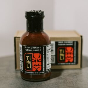 A bottle of Ti Mo Li Indo-Chinese Fusion Sauce with a black cap stands in front of a box featuring the same label, on a gray surface with a plain background.