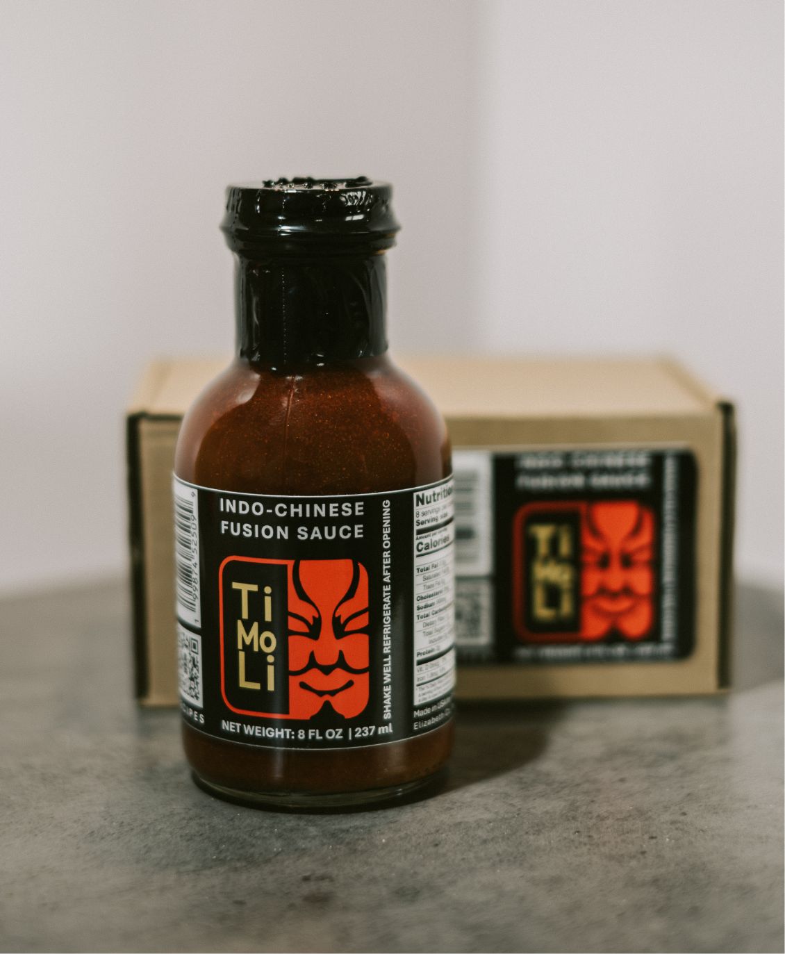 A bottle of Ti Mo Li Indo-Chinese Fusion Sauce with a black cap stands in front of a box featuring the same label, on a gray surface with a plain background.