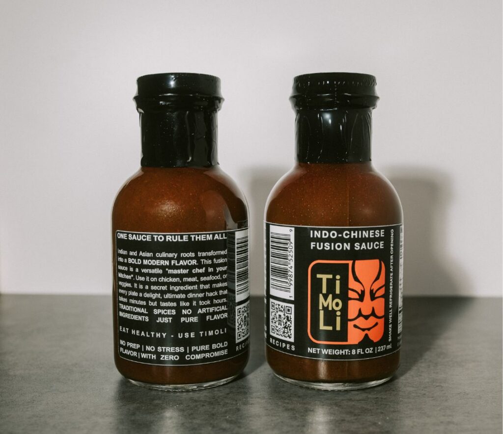 Two glass bottles of Indo-Chinese fusion sauce labeled TiMoLi sit side by side on a gray surface, with one bottle showing the front label and the other showing the back label with ingredient and flavor descriptions.